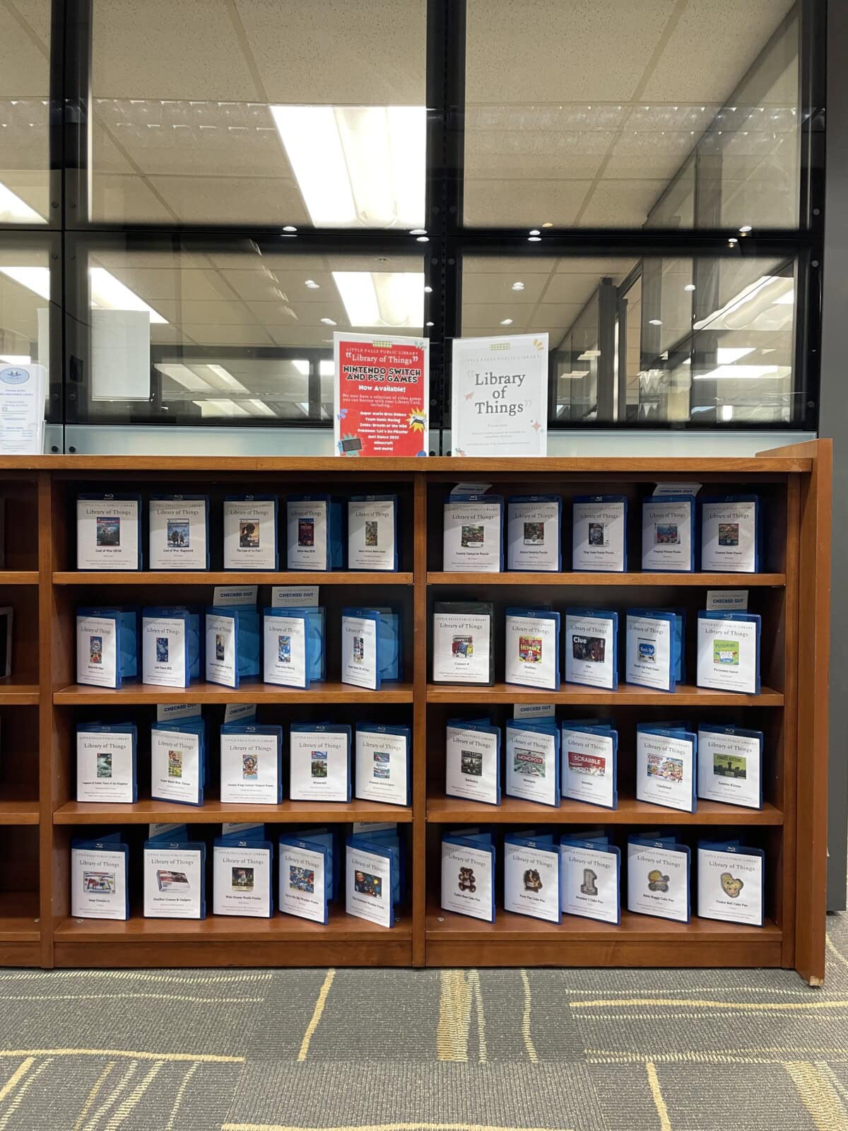 A wooden bookshelf displays labeled cards for various items available to borrow, with a sign on top reading "Library of Things" in a library setting.
