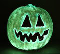 A glowing jack-o'-lantern with triangular eyes and a jagged mouth carved into its surface, illuminated in green against a dark background.